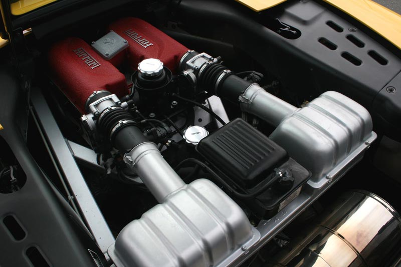 Blick in den Motorraum eines gelben Ferrari mit V8-Motor, roten Ansaugbrücken mit Ferrari-Schriftzug und silbernen Luftfiltergehäusen