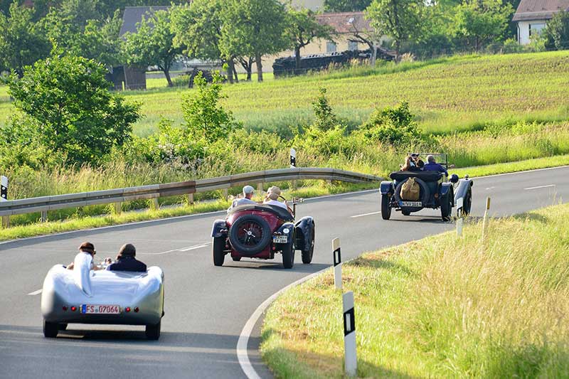Drei offene Oldtimer, darunter ein silberner Rennsportwagen, fahren hintereinander auf einer kurvigen Landstraße zwischen Wiesen