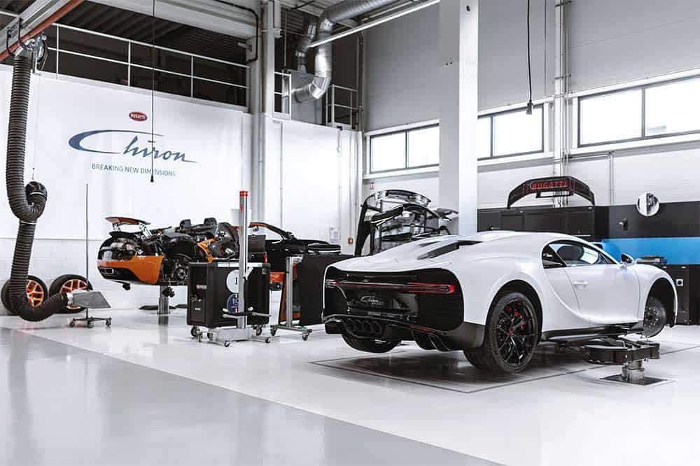 Bugatti-Werkstatt mit einem weißen Chiron auf der Hebebühne, daneben ein zerlegter orange-schwarzer Bugatti und Chiron-Banner