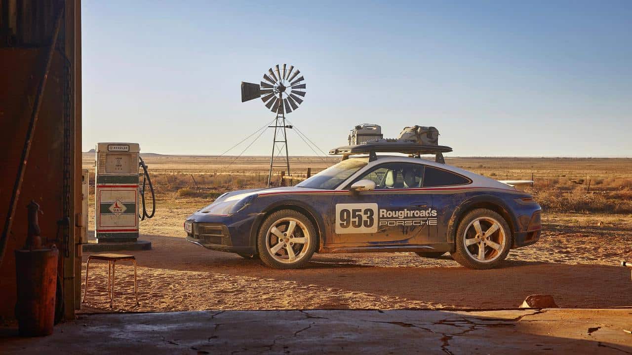Blau-weißer Porsche 911 Dakar mit Startnummer 953 und Dachgepäck neben einer alten Zapfsäule und Windrad in karger Steppe