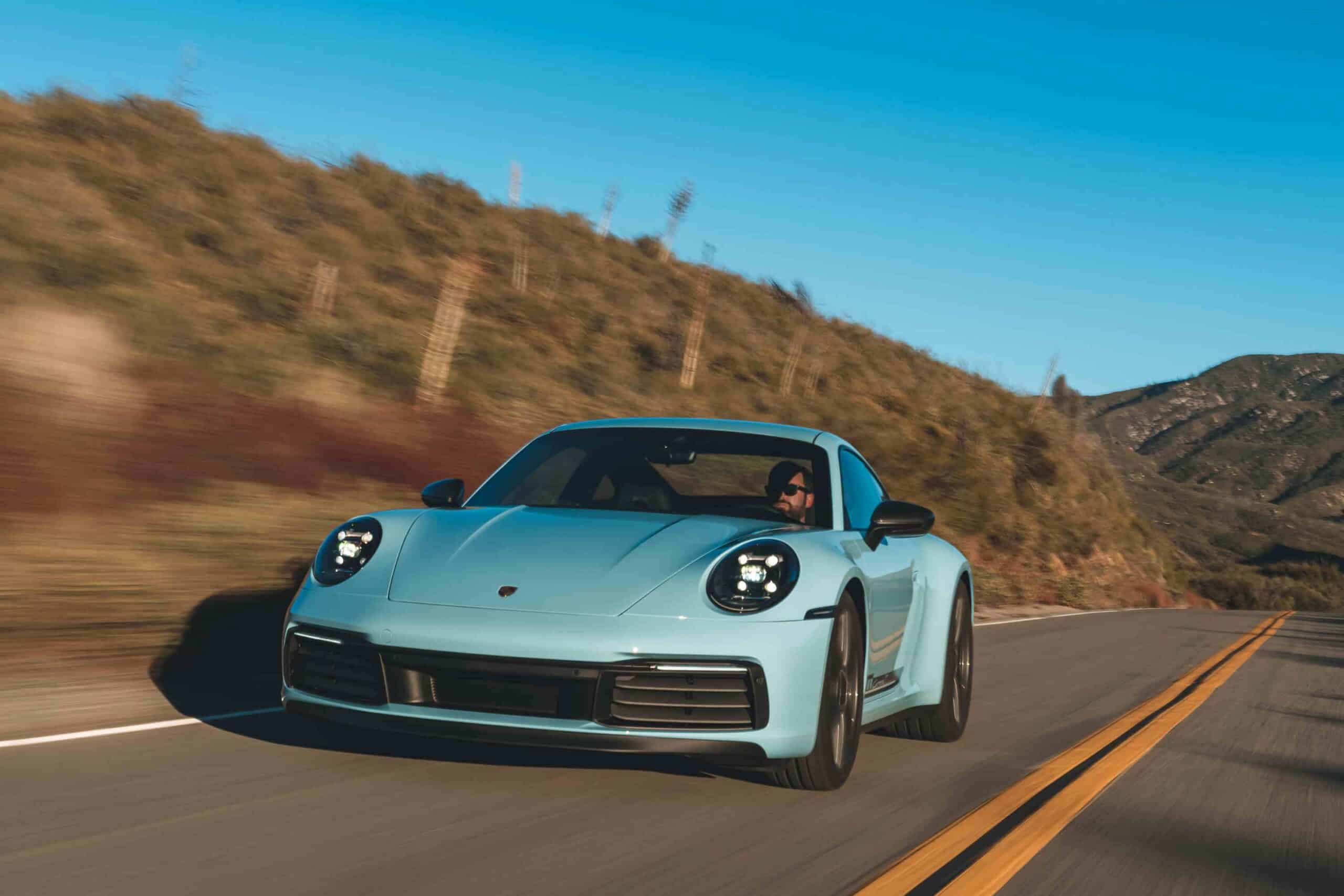 Hellblauer Porsche 911 Carrera T in Fahrt auf einer kurvigen Landstraße vor trockenen Hügeln und blauem Himmel