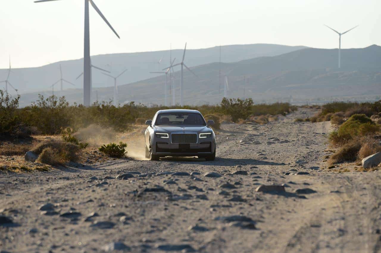 Grauer Rolls-Royce Ghost fährt auf einer staubigen Schotterpiste durch Buschland, dahinter Windräder und Berge