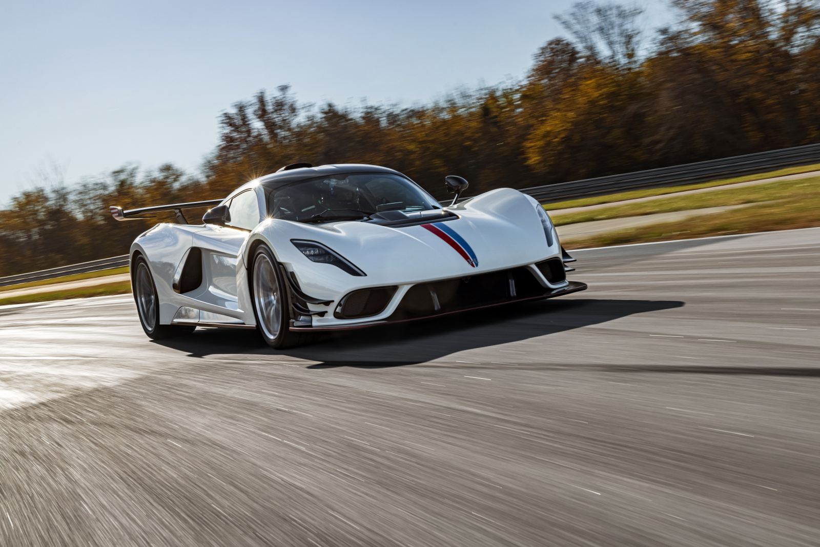 Weißer Hennessey Venom F5 mit rot-blauem Streifen auf der Haube fährt über eine Rennstrecke, im Hintergrund Herbstbäume
