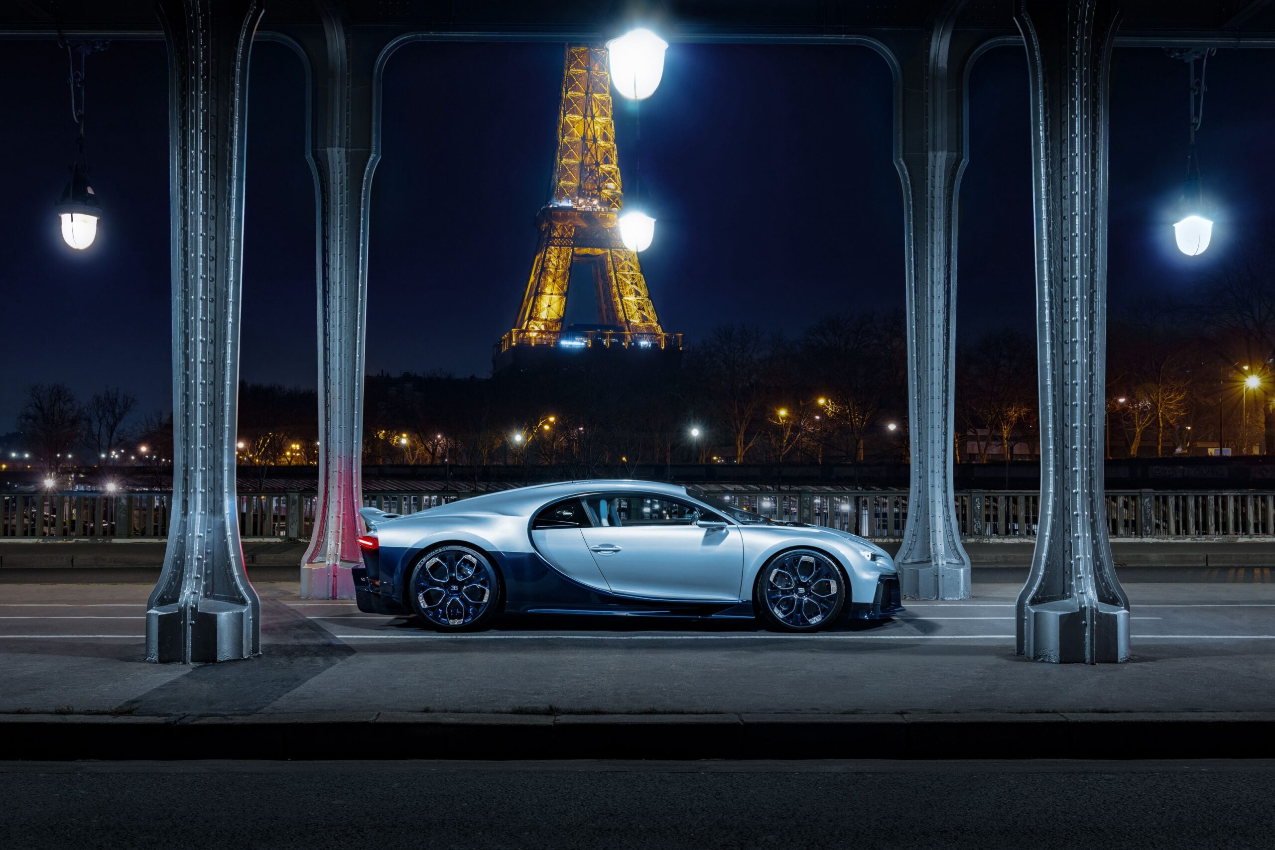 Silber-blauer Bugatti Chiron Profilée in Seitenansicht unter einer beleuchteten Brücke, dahinter der nächtlich angestrahlte Eiffelturm